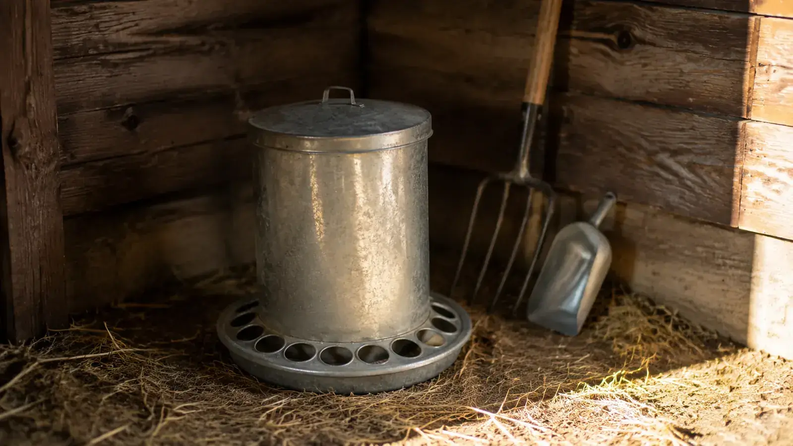 Grandpa's Feeders galvanized treadle chicken feeder with the lid closed, sitting in a straw-lined coop corner with a tool leaning against the wall